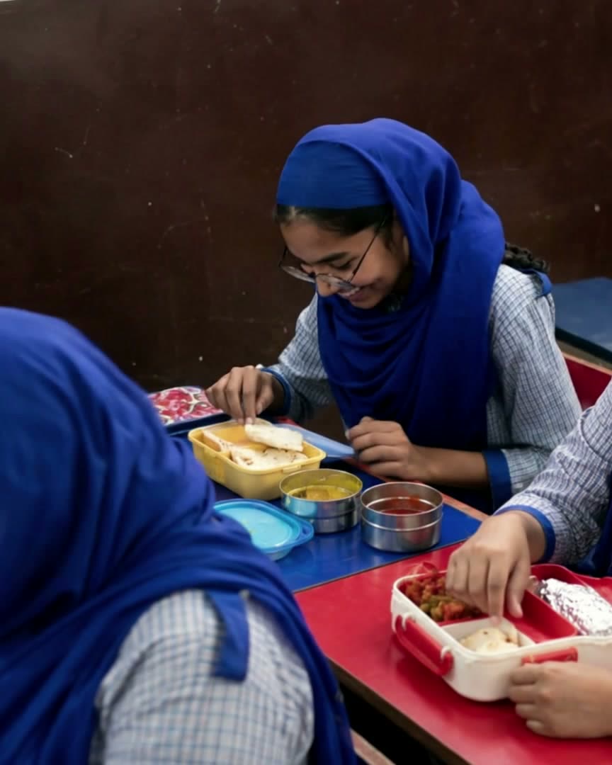 Four schoolgirls in blue hijabs share a lunch tiffin together, laughing.